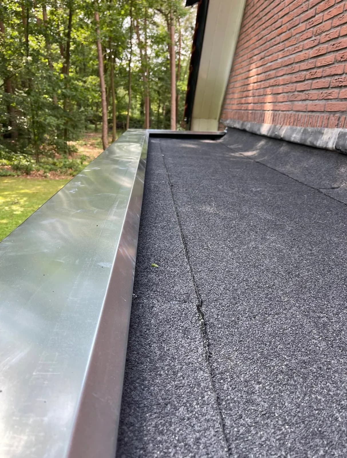 Nieuw zwart bitumen plat dak met zinken daktrim langs een bakstenen gevel, bosrand op de achtergrond
