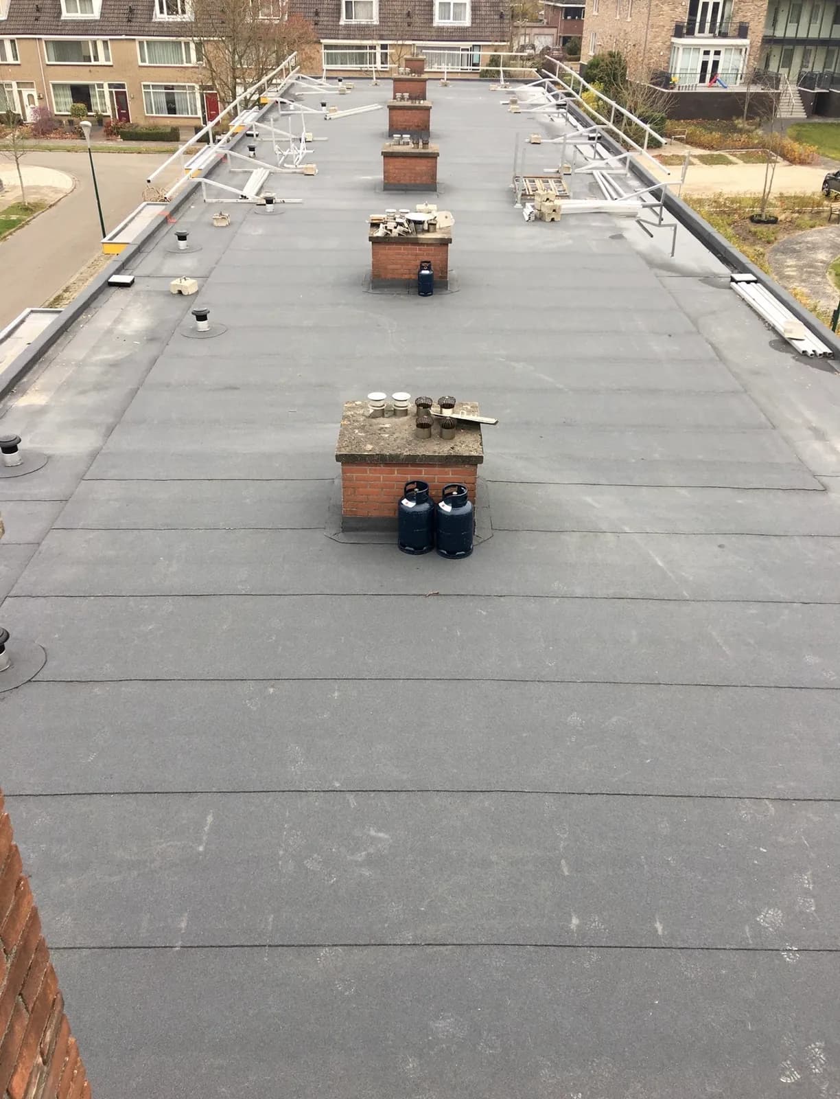 Groot plat dak van een flat met nieuw grijs bitumen, meerdere gemetselde schoorstenen en dakrandbeveiliging, gezien van bovenaf